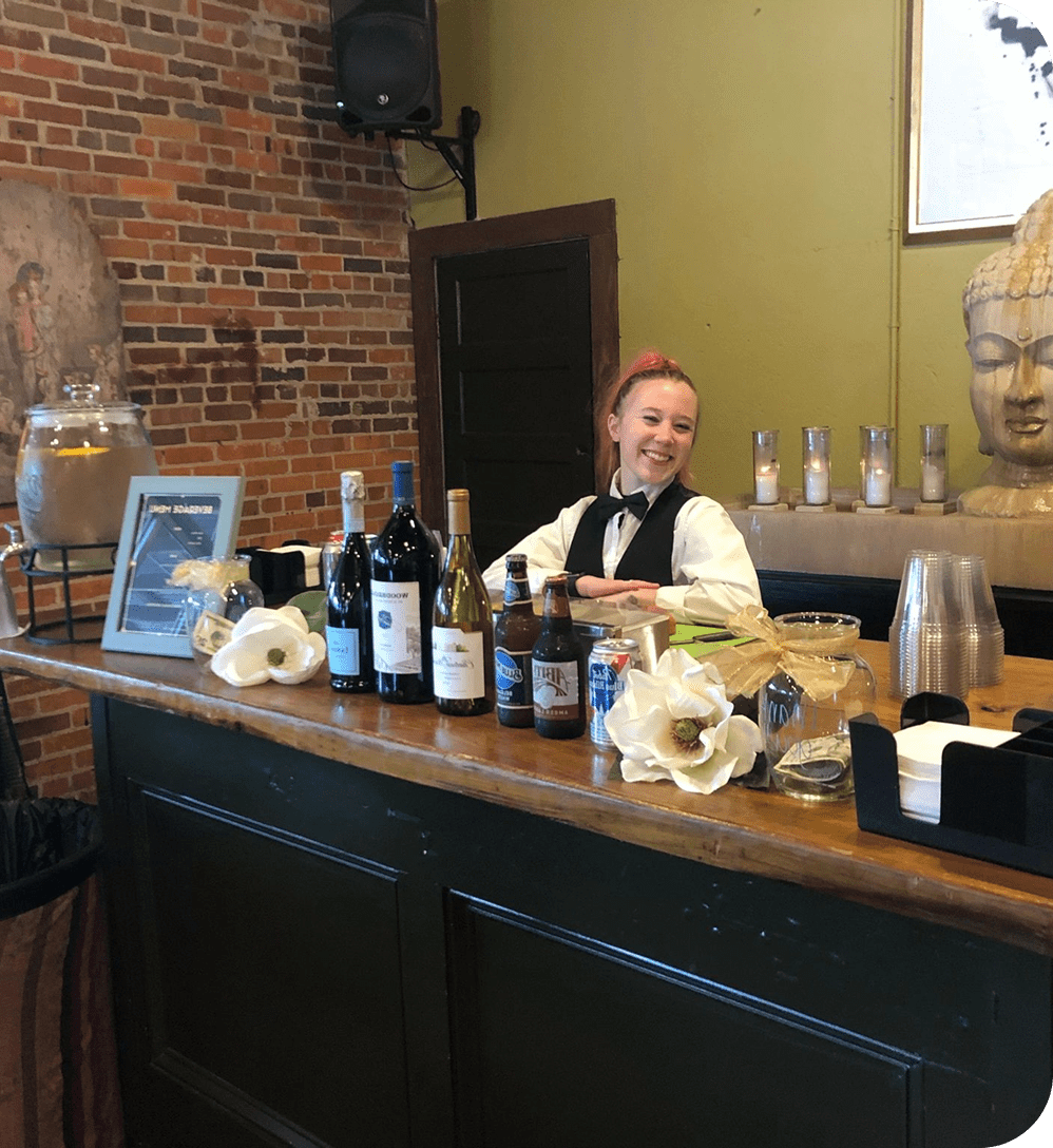 Smiling bartender behind bar with drinks displayed.